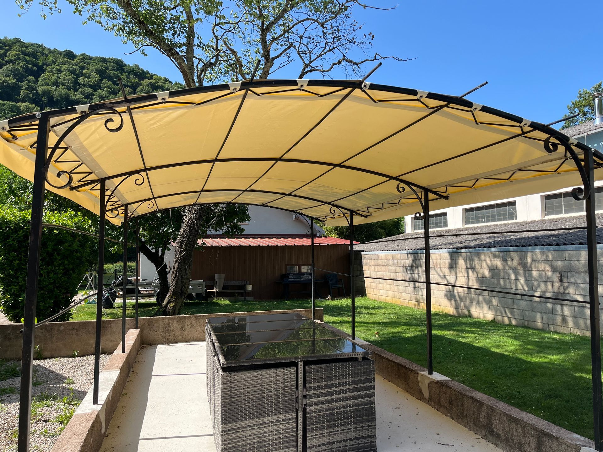 Un auvent beige recouvre une table sur une terrasse. Herbe verte et arbres en arrière-plan. Ciel bleu.