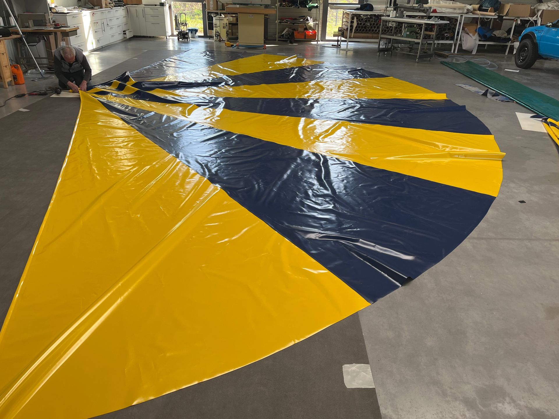 Un grand tissu à rayures jaunes et bleu marine est étalé sur un sol gris. Une personne travaille sur le bord.