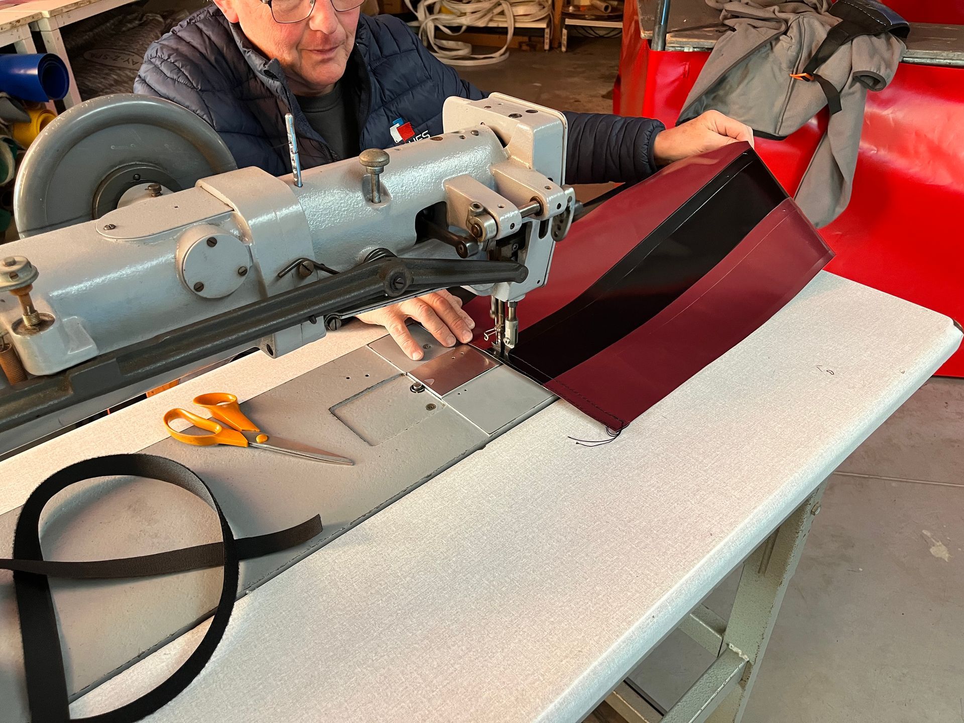Une personne coud du tissu rouge et noir sur une machine à coudre industrielle, dans un atelier.