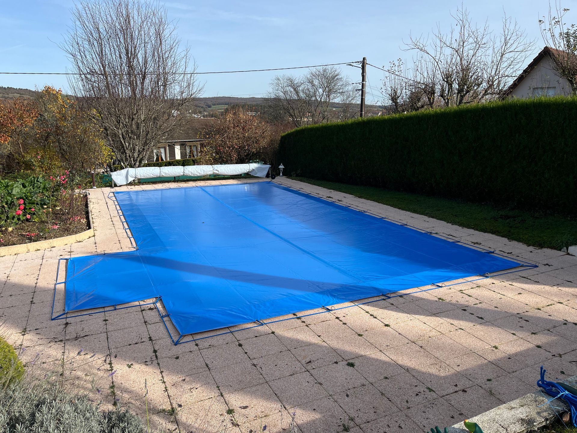 Couverture de piscine bleue sur une piscine rectangulaire dans un jardin, entourée de pierres et de verdure.