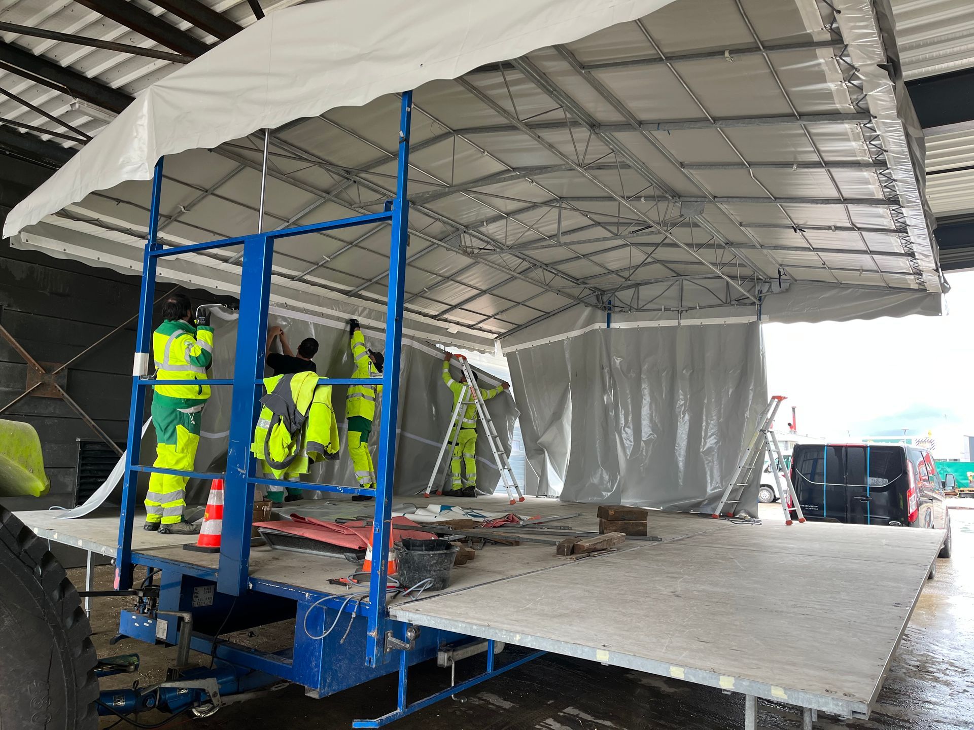 Des ouvriers portant des gilets de sécurité assemblent un abri temporaire sur une remorque à plateau.
