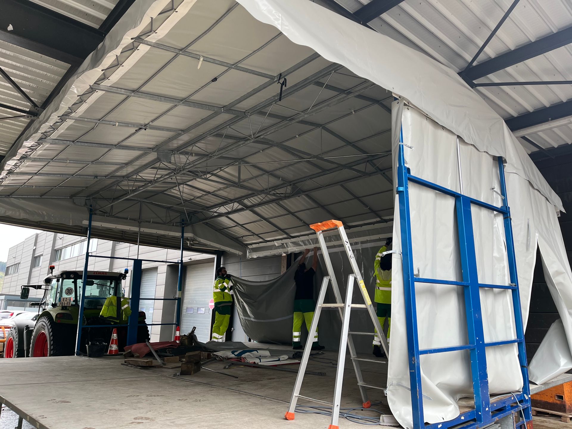 Des ouvriers installent une structure de tente blanche, un tracteur et une échelle sont présents.