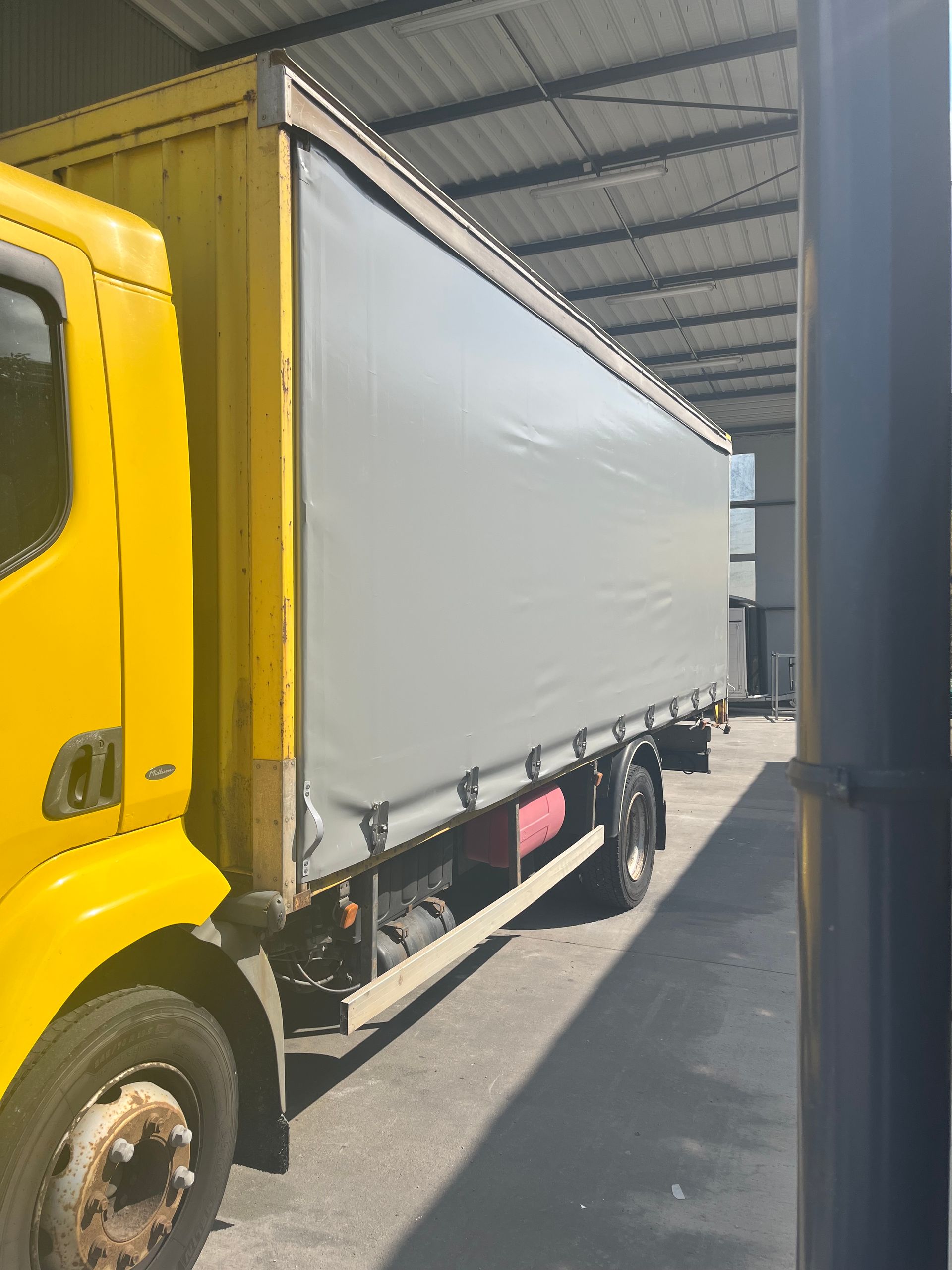 Camion de livraison jaune avec une zone de chargement recouverte d'une bâche grise, stationné sous un toit métallique.