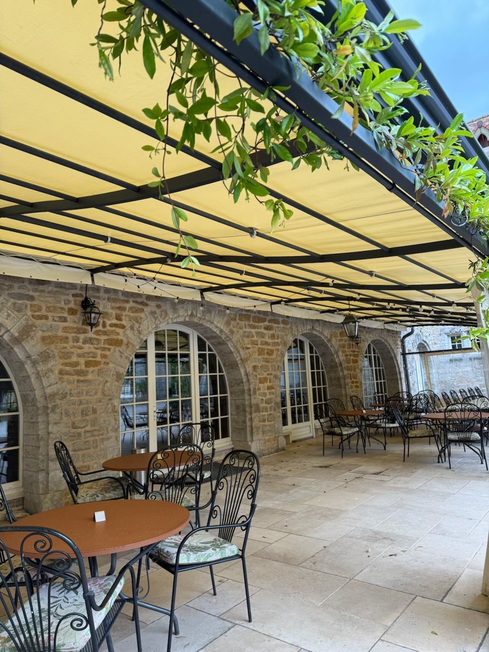 Terrasse extérieure avec mur en pierre, fenêtres cintrées, tables, chaises et auvent jaune.