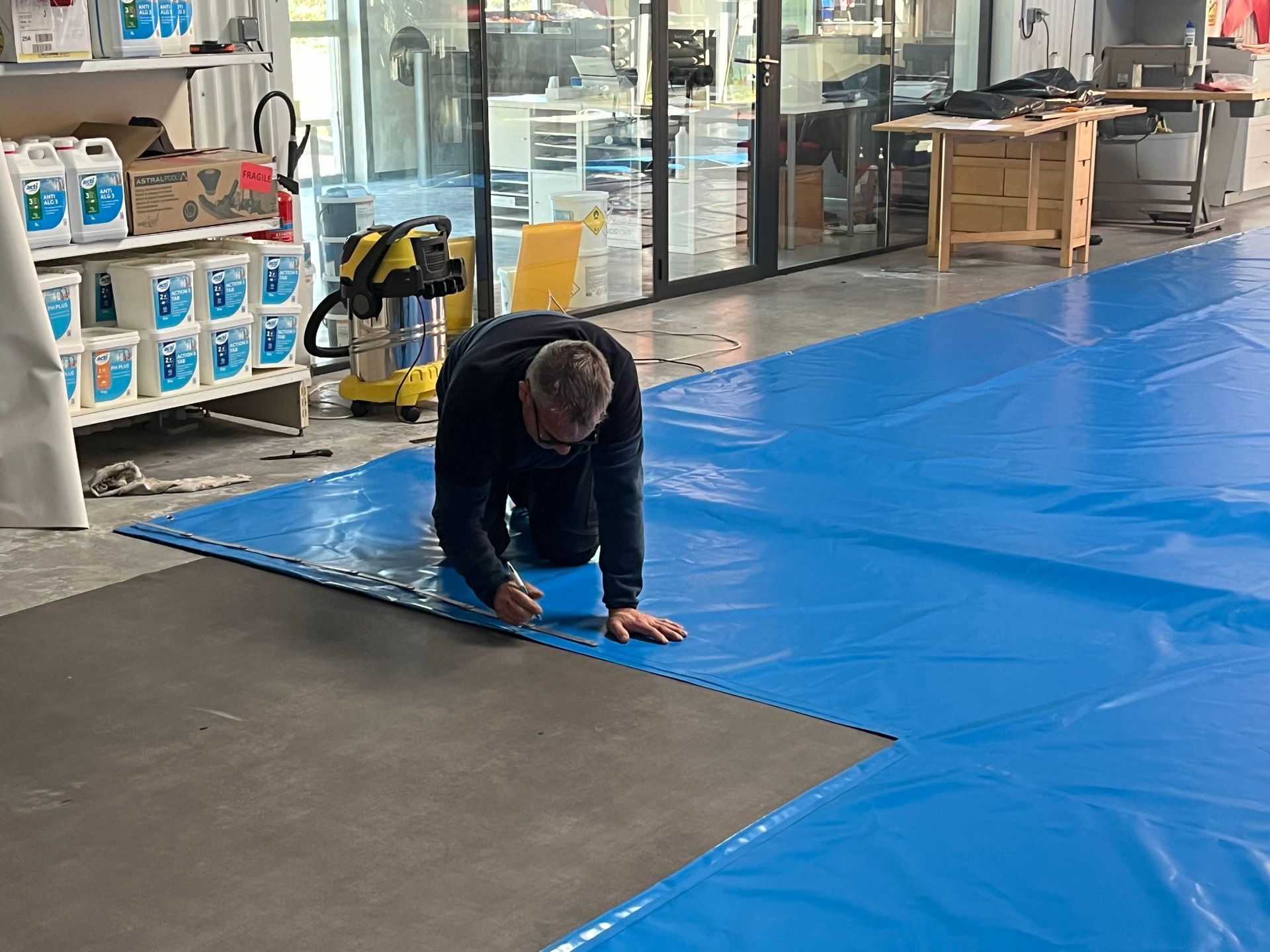 Une personne agenouillée trace des lignes à la règle sur une bâche bleue dans un entrepôt.  