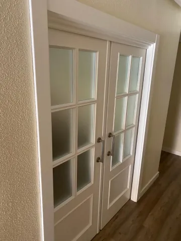 Puertas dobles blancas de estilo francés con paneles de vidrio en un pasillo, junto a un suelo de madera y paredes beige