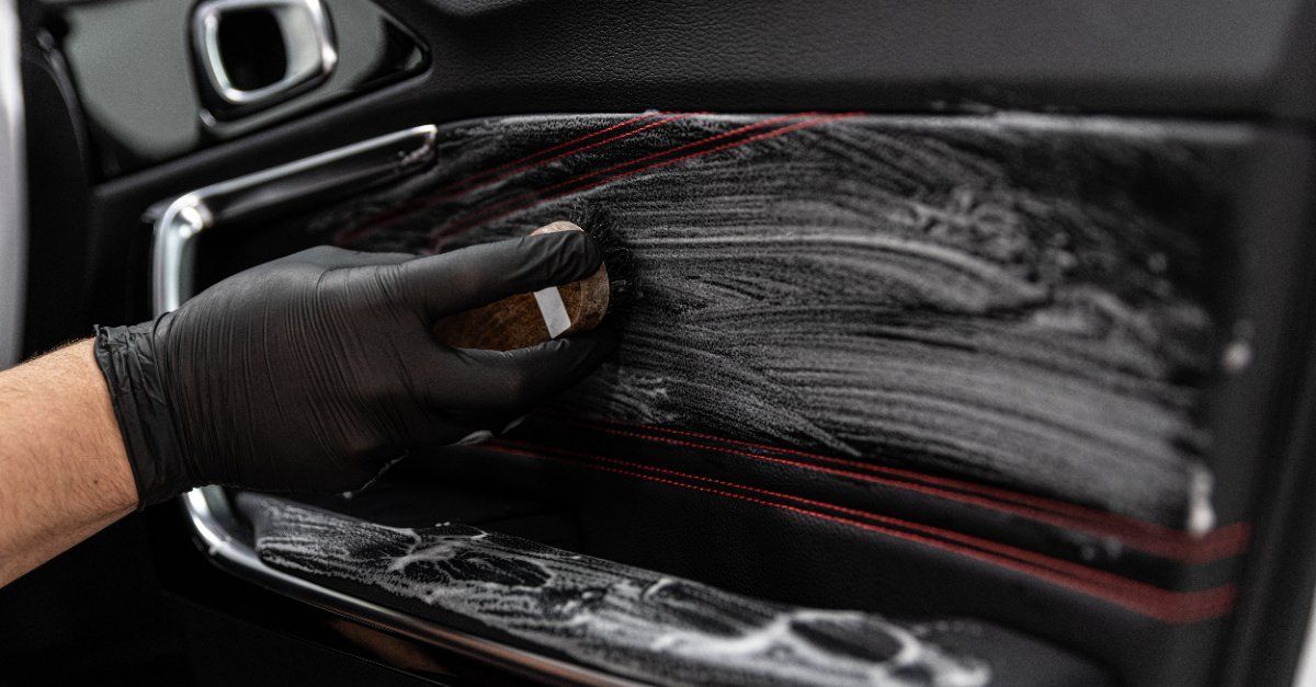 Una persona que lleva un guante negro frota el panel de la puerta de un automóvil con un cepillo y una solución limpiadora.