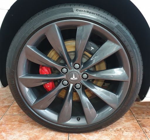 Primer plano de una rueda Tesla con llanta gris, neumático negro y pinza de freno roja. El coche se encuentra en un entorno de azulejos.