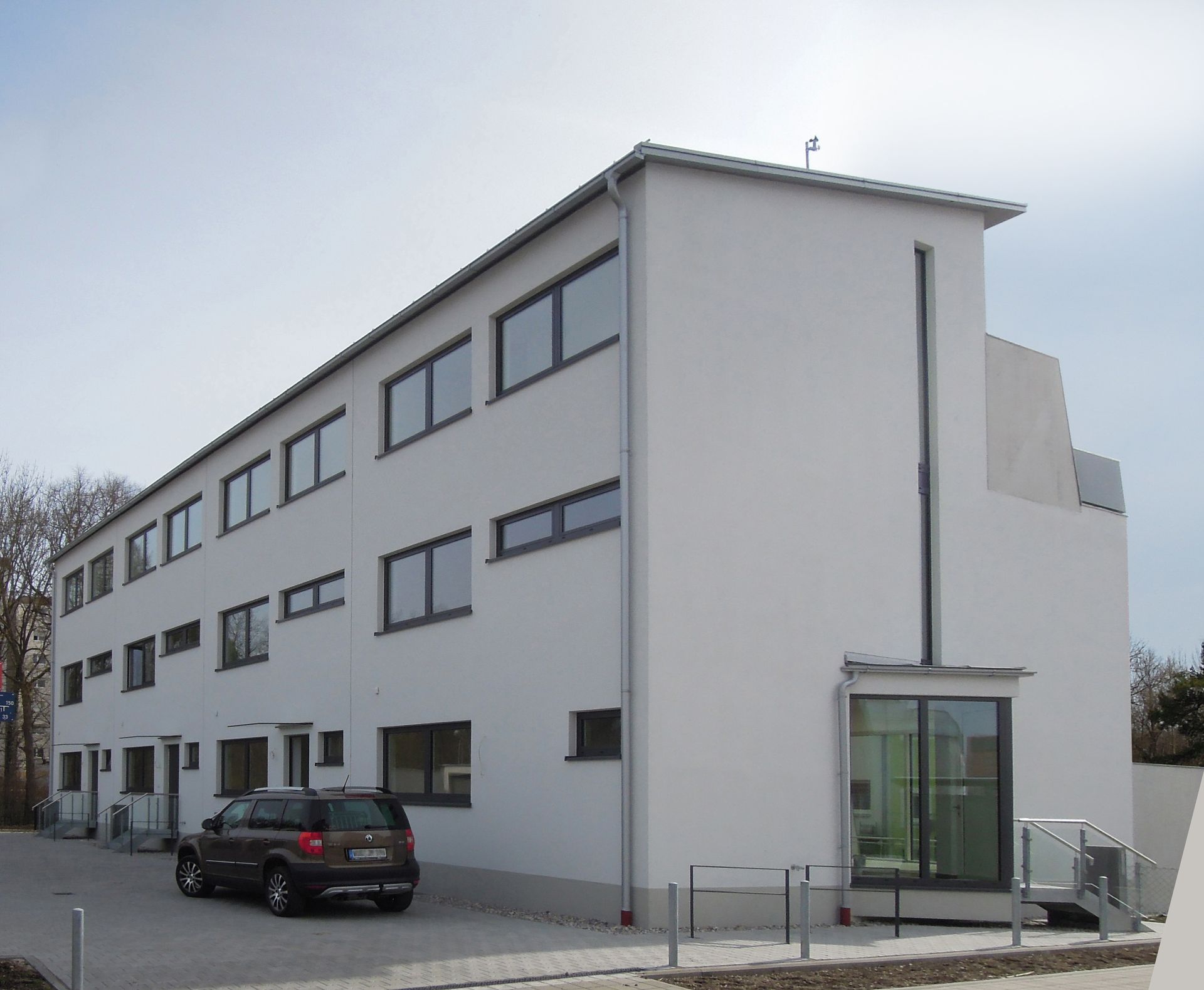 Weißes, dreistöckiges Gebäude mit Fensterreihen, einem kleinen Glaseingang und einem davor geparkten Auto.