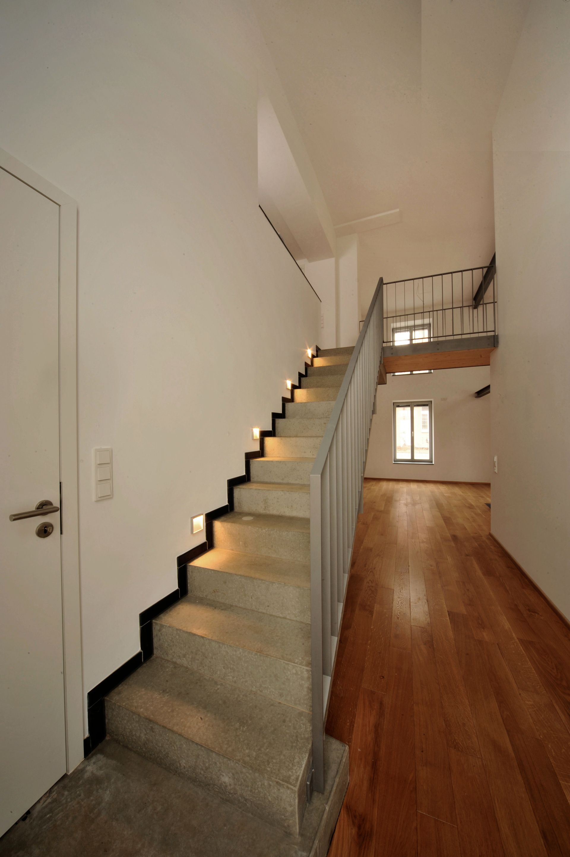 Eine Treppe mit eingebauter Beleuchtung führt zu einem kleinen Balkon mit Metallgeländer, Holzböden und weißen Wänden.