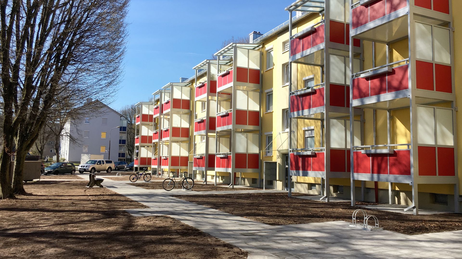 Mehrfamilienhaus mit rot-weißen Balkonen, gelben Wänden und einem von Bäumen gesäumten Gehweg an einem sonnigen Tag.