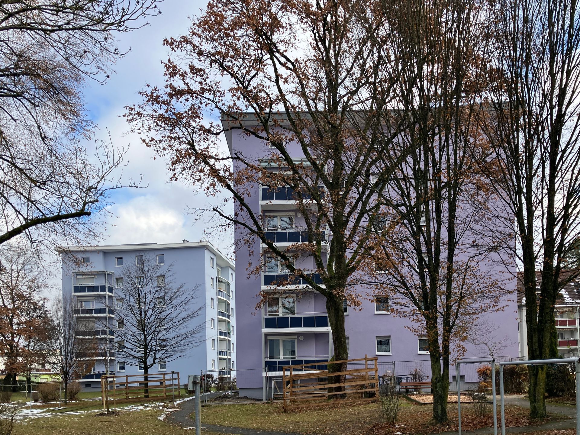 Zwei hellviolette Wohnhäuser mit Balkonen, kahlen Bäumen und einem umzäunten Spielbereich an einem bewölkten Tag.