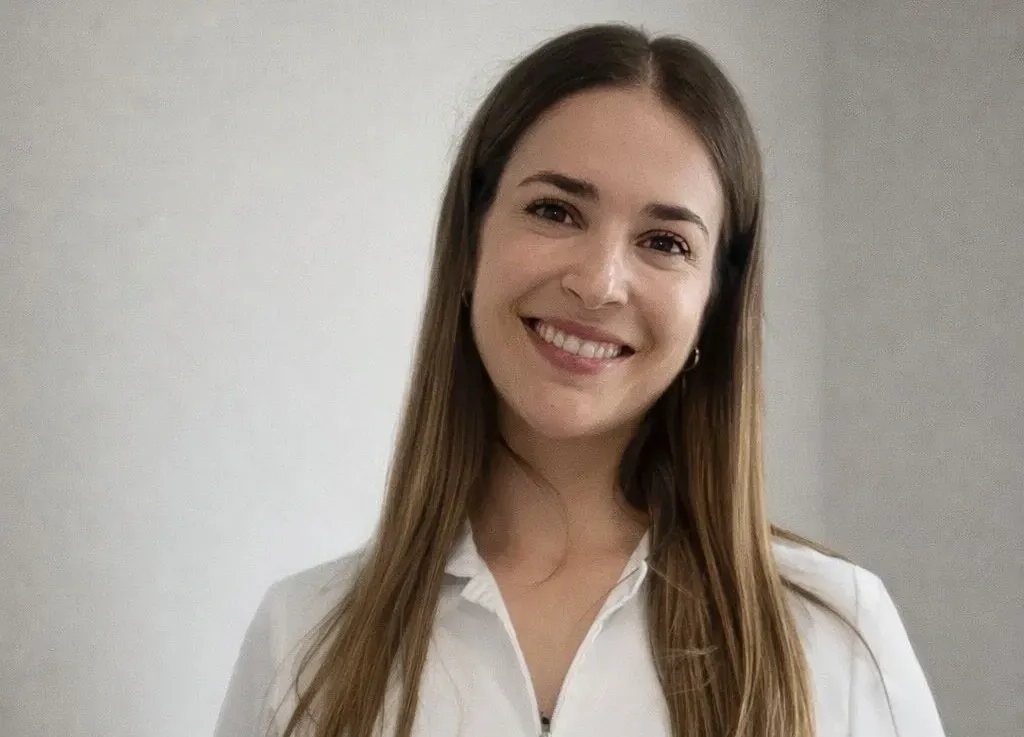 Una persona sonriente con cabello largo, liso y castaño lleva una camisa blanca con cuello sobre un fondo gris neutro.