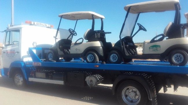Tres carros de golf en una grúa azul, estacionados en un día soleado.