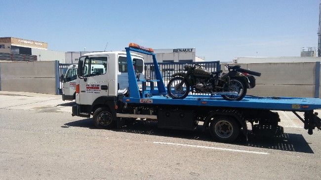 Grúa con una motocicleta negra en su plataforma, estacionada sobre la acera en un día soleado.