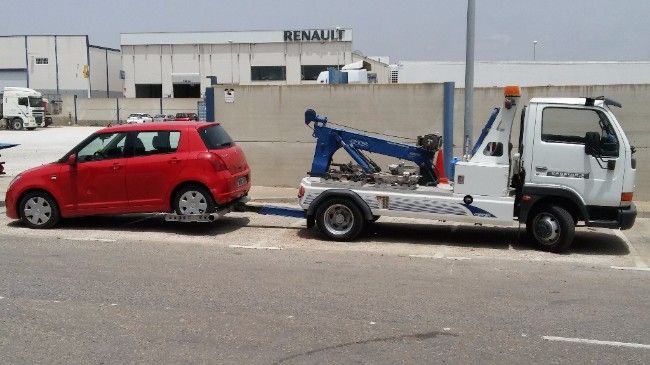 Un automóvil rojo es remolcado por una grúa blanca, estacionado en una calle cerca de un edificio Renault.