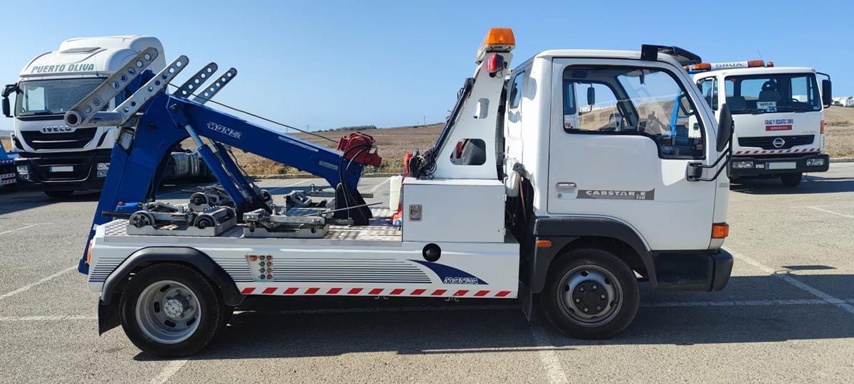 Grúa blanca con pluma azul, estacionada sobre una superficie pavimentada, con otros camiones al fondo. Día soleado.