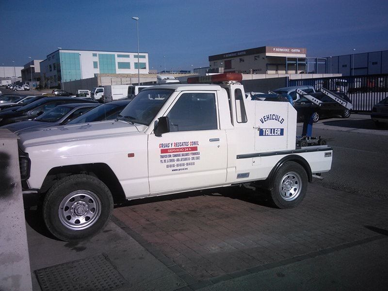 Vehículo de servicio blanco estacionado en un aparcamiento, con un texto en el lateral. Un día soleado y brillante, con otros coches y edificios al fondo.