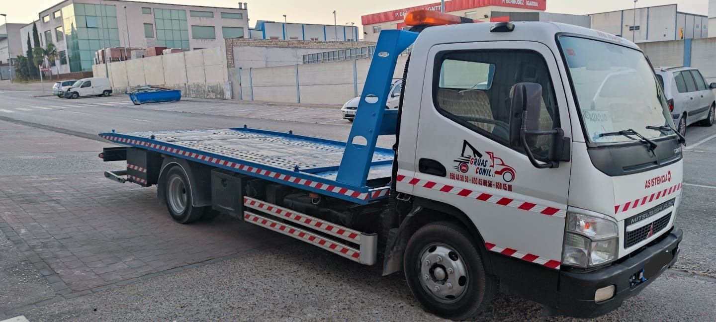 Una grúa blanca con plataforma, rampa azul y luces naranjas estacionada en la acera.