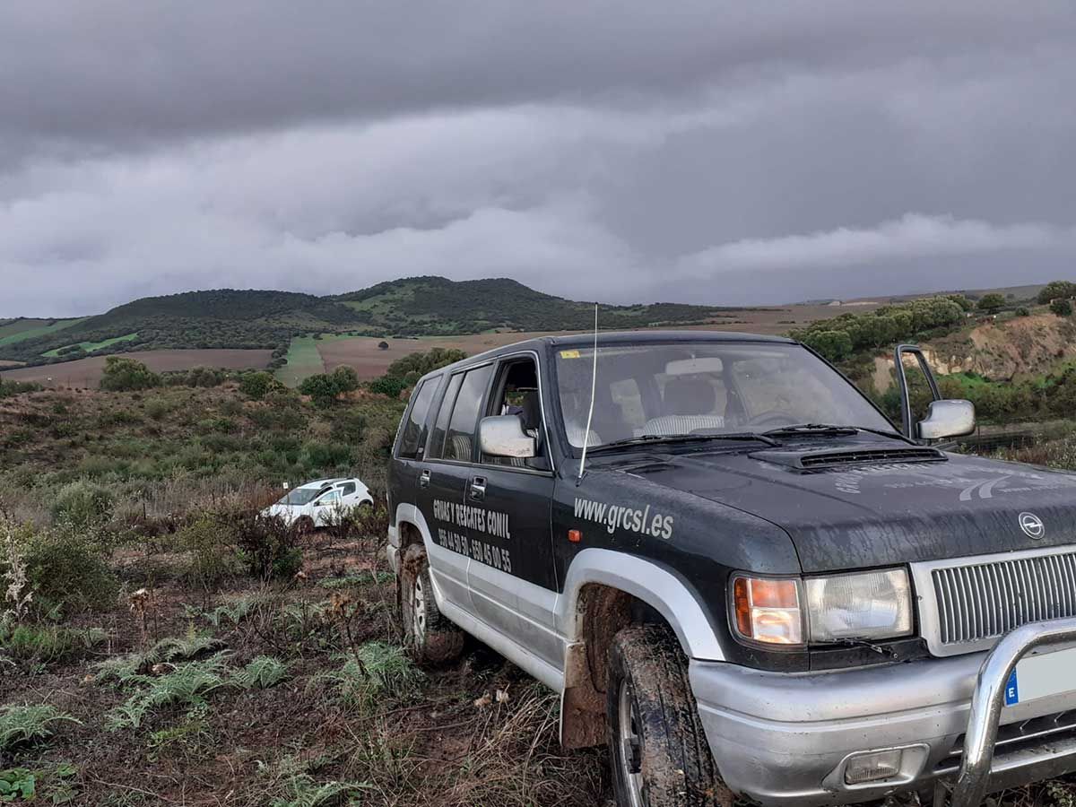 SUV oscuro sobre terreno fangoso con un auto blanco a lo lejos, colinas y un cielo nublado.
