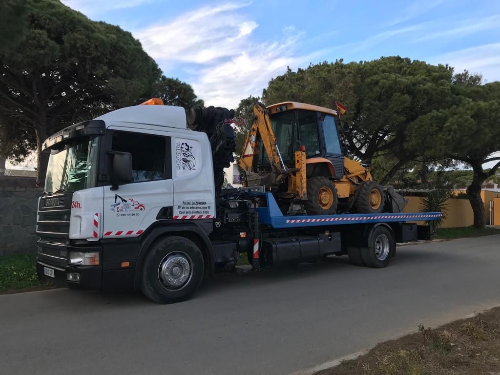 Grúa que transporta una retroexcavadora amarilla en una carretera, árboles en el fondo.