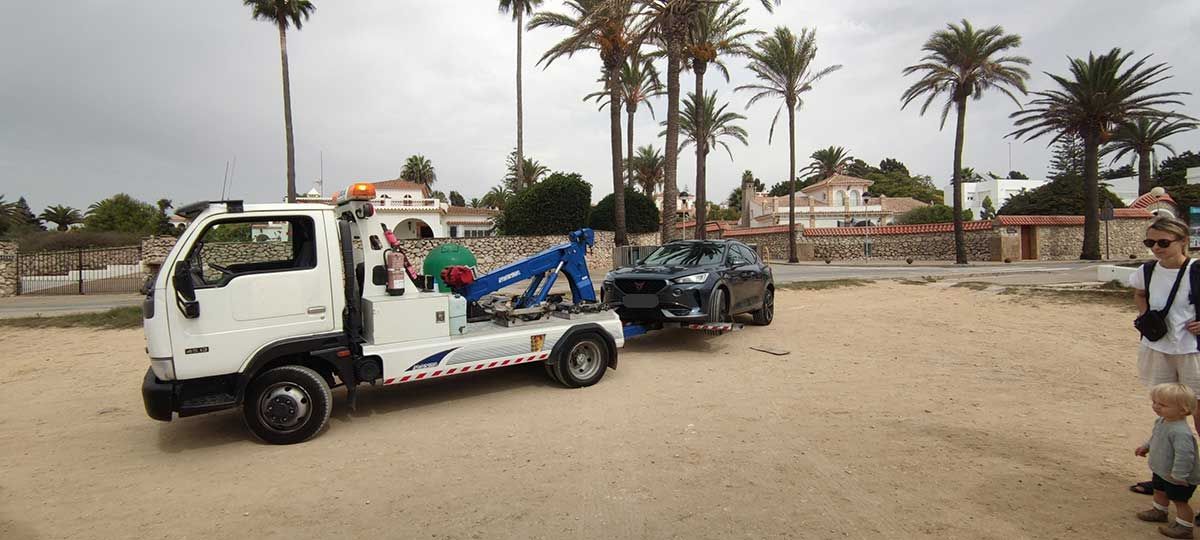 Una grúa blanca remolca un coche oscuro por una zona arenosa, con palmeras y edificios al fondo.