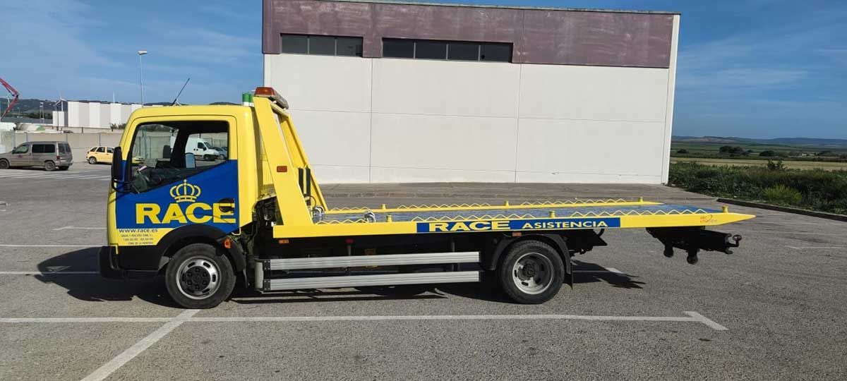 Grúa RACE amarilla y azul en un lote pavimentado, con un edificio blanco y marrón al fondo.