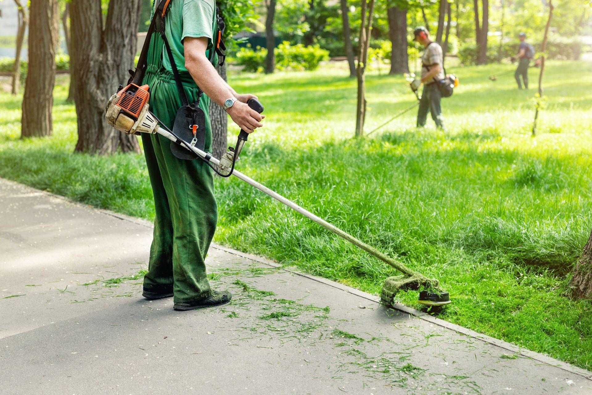 Un homme en salopette verte utilise une débroussailleuse pour tailler l'herbe le long d'un trottoir dans un parc.