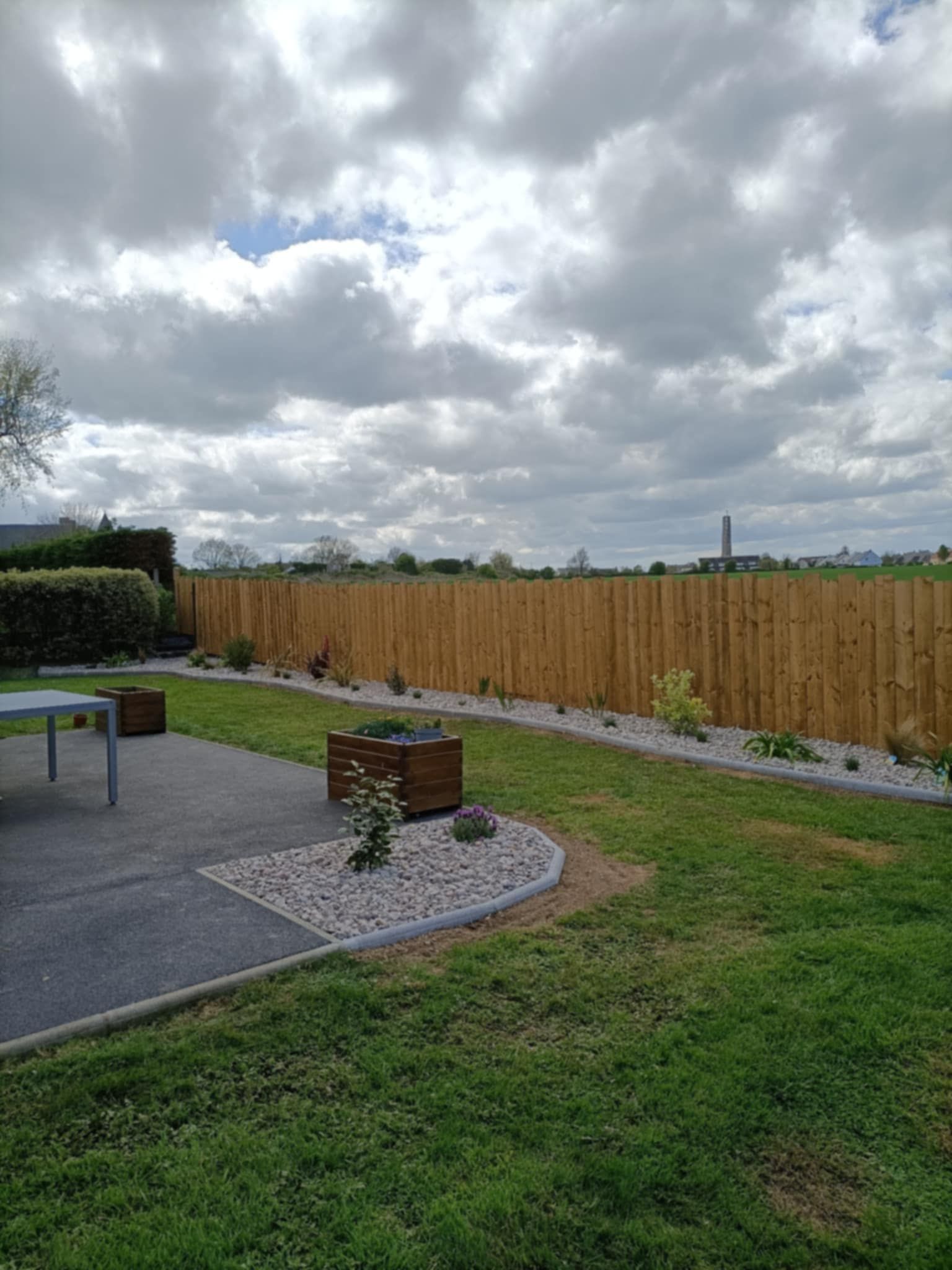 Pelouse et jardin d'une maison moderne ; terrasse avec table et chaises. Haie de conifères à l'arrière.