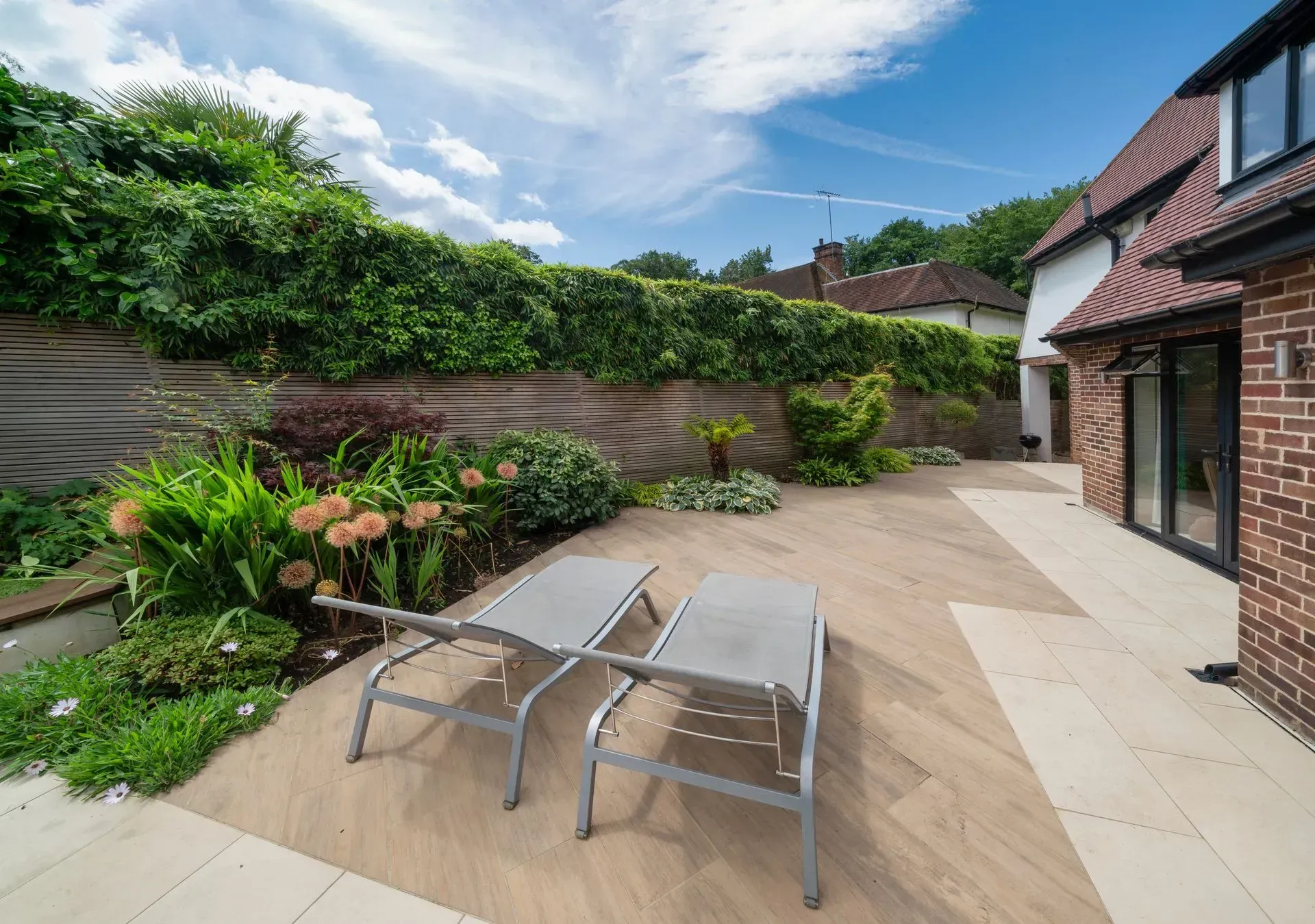 Deux chaises longues sur une terrasse donnant sur un jardin et une maison en briques. Verdure luxuriante et ciel bleu.