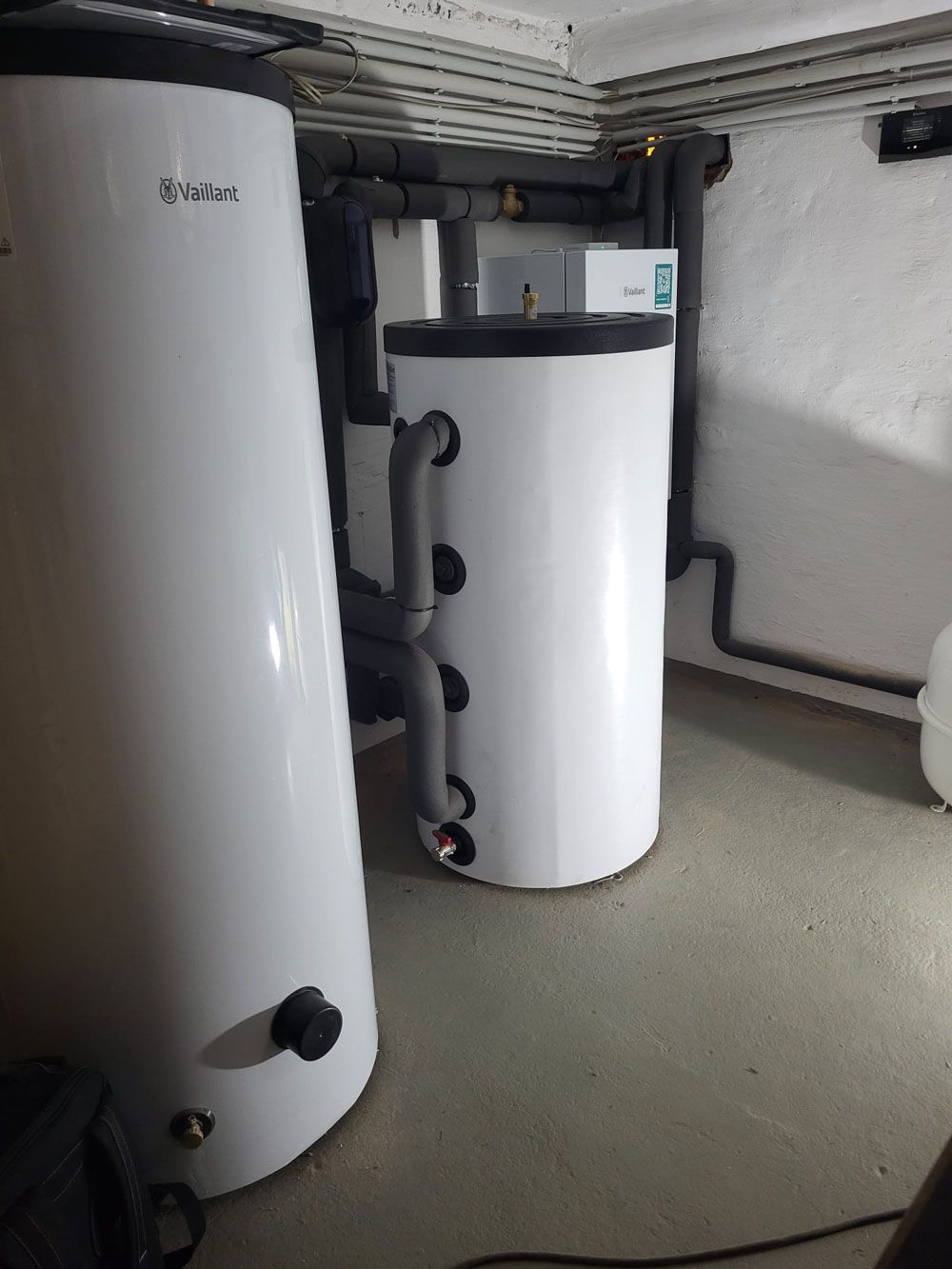 Weiße, zylindrische Warmwasserbereiter in einem Keller oder Hauswirtschaftsraum, mit freiliegenden Rohren und Deckenbalken.