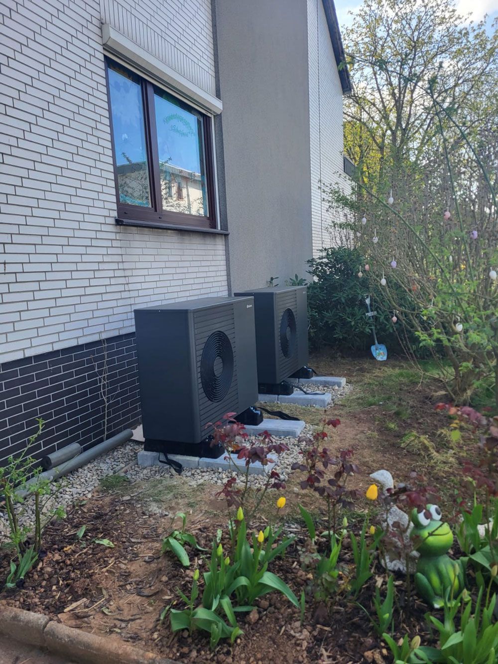 Zwei Außenklimaanlagen neben einem weißen Backsteinhaus, im Vordergrund ein Gartenbeet mit Blumen.