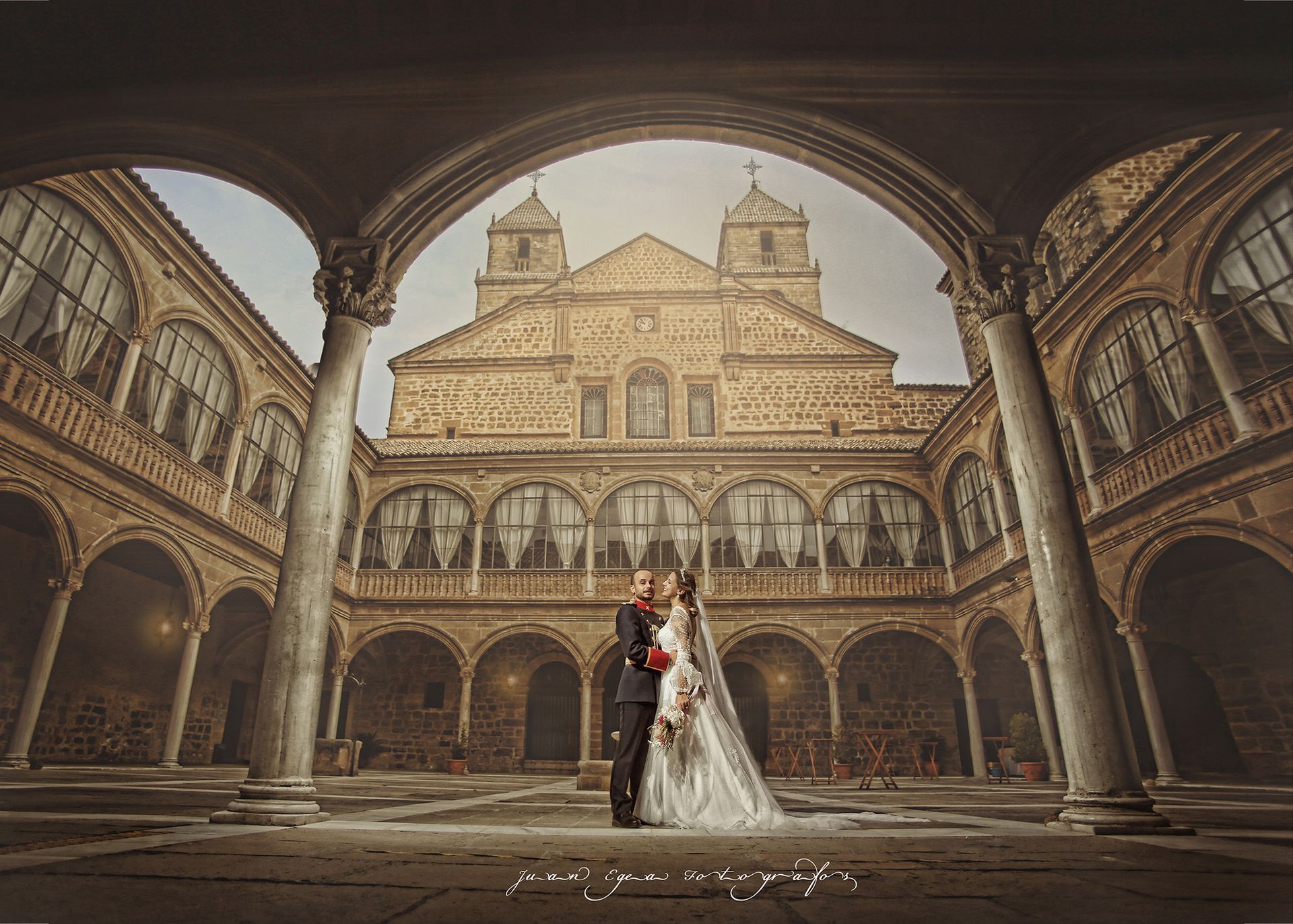 Pareja de novios abrazándose en un patio histórico con columnas arqueadas y un edificio ornamentado como telón de fondo.