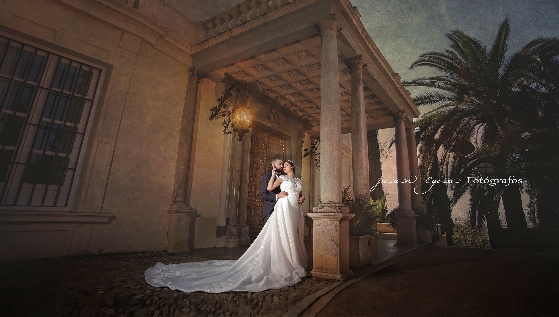 Los novios se abrazan bajo un porche con columnas con un vestido blanco suelto y una palmera al fondo.