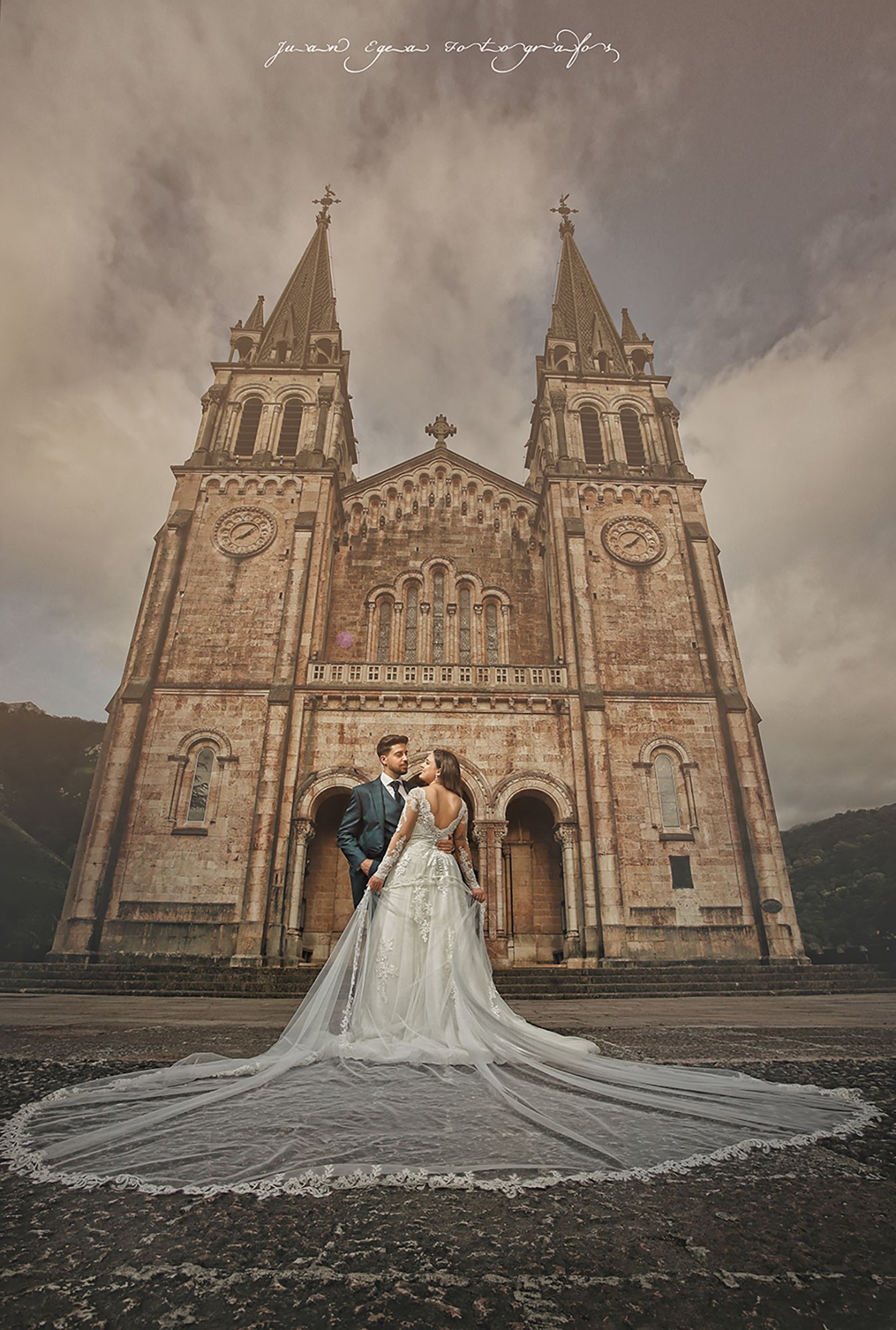 Pareja besándose frente a una catedral. La novia con vestido blanco y larga cola; el novio con traje azul marino. Cielo nublado.