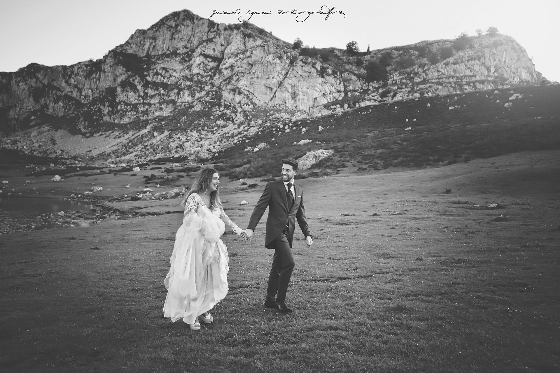 Los novios se dan la mano, caminando por una llanura con montañas de fondo. Foto en blanco y negro.