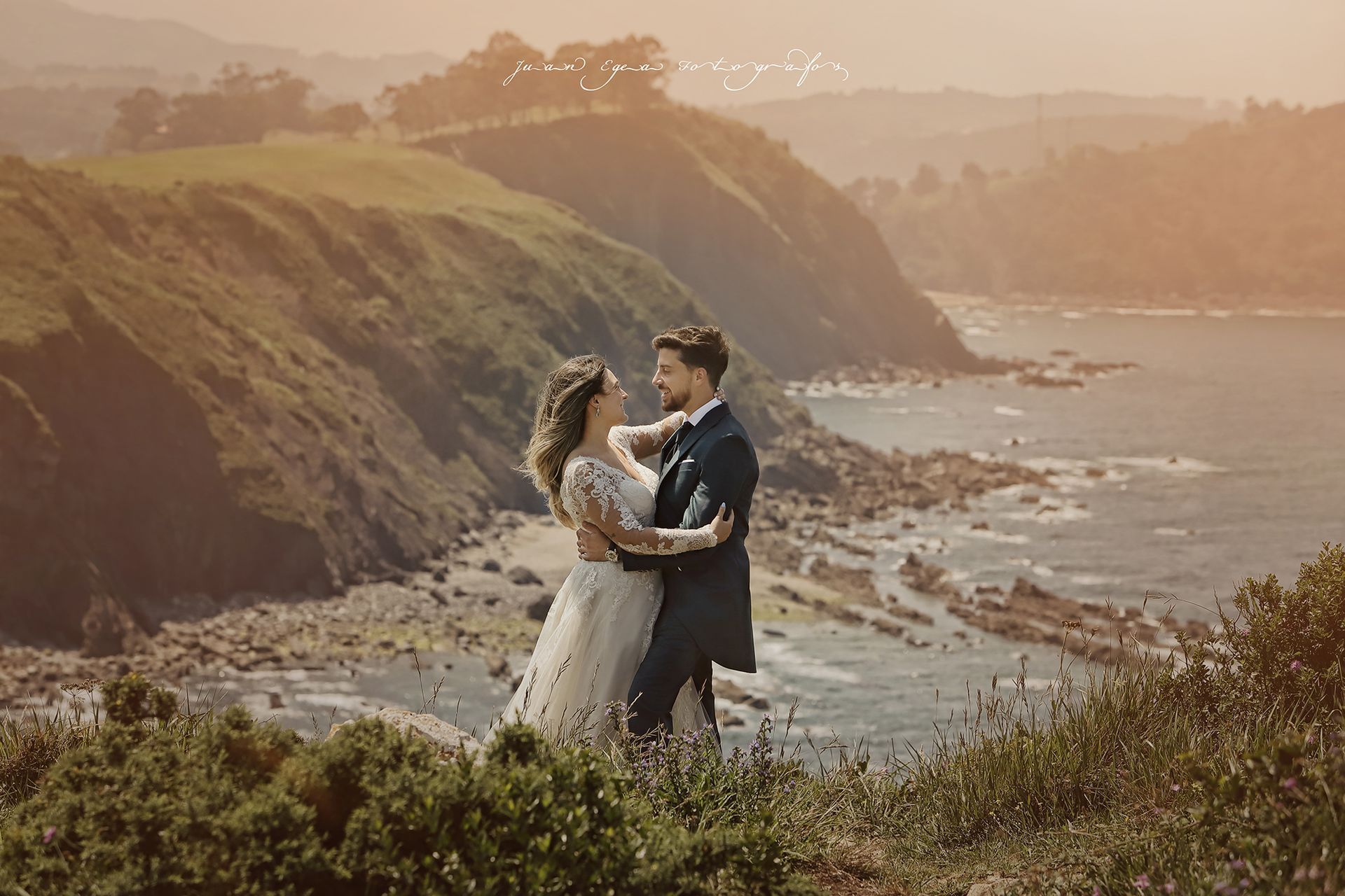 Los novios se abrazan en un acantilado con vistas al océano. La suave luz del sol baña la escena.