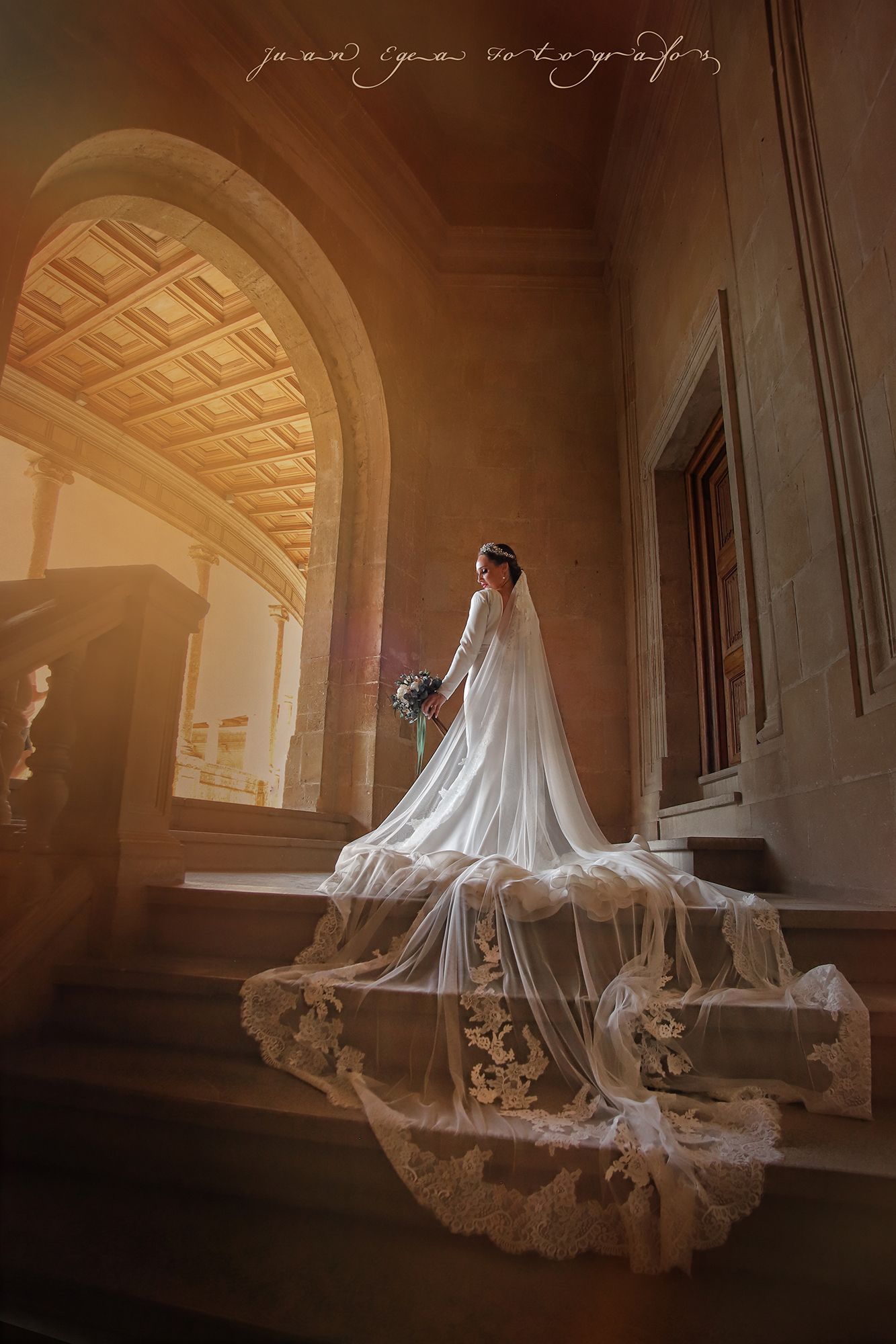 Novia con vestido blanco y larga cola sobre escalones de piedra, con arco iluminado por el sol como fondo.
