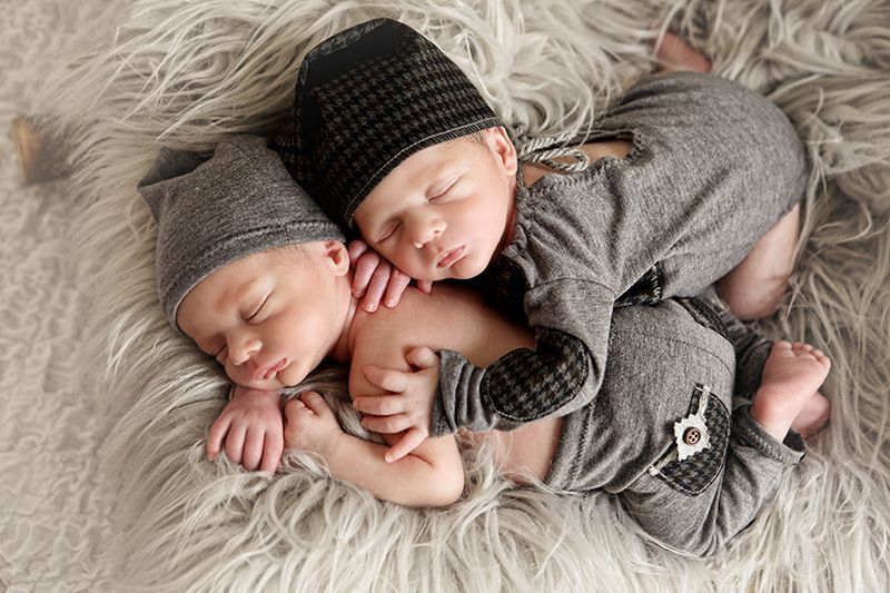 Dos bebés recién nacidos durmiendo, vestidos con trajes y sombreros grises, acurrucados sobre un pelaje gris y esponjoso.