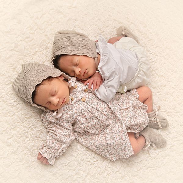 Dos bebés recién nacidos durmiendo, con gorros y trajes florales, sobre una manta de color crema.