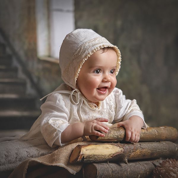 Bebé sonriente con gorro y vestido blancos, sosteniendo troncos, en un entorno rústico.