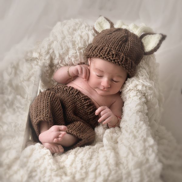 Bebé durmiendo, vestido con un sombrero de ciervo marrón y pantalones cortos, acurrucado sobre una manta blanca y esponjosa.