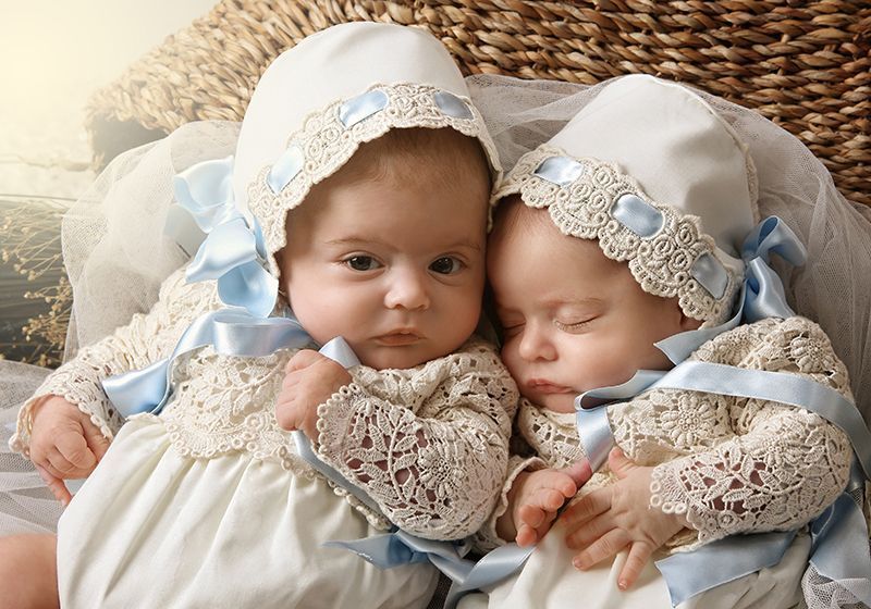 Dos bebés con trajes de encaje blanco y gorros, uno despierto y mirando al espectador, el otro dormido.