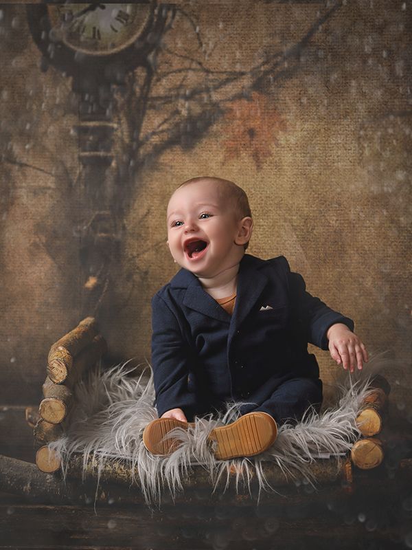 Un bebé con traje azul marino se ríe mientras está sentado sobre una piel, sobre un fondo rústico con un reloj.