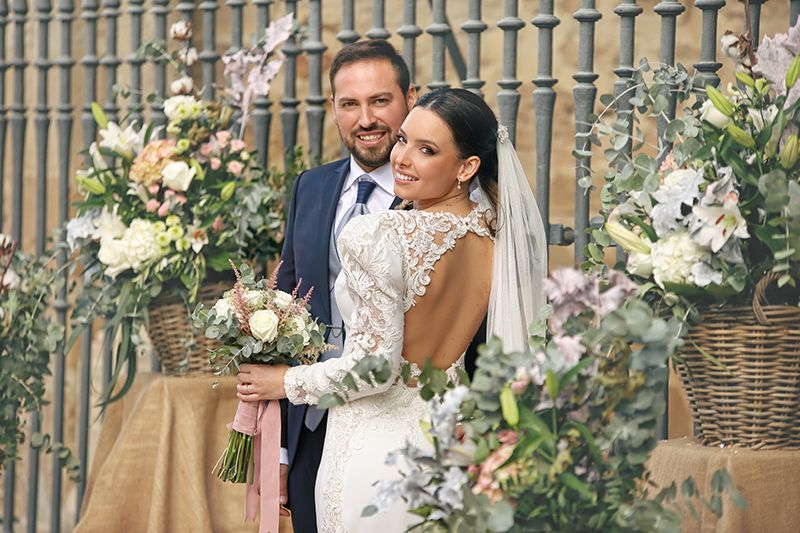 Pareja de novios sonriendo. La novia con vestido de encaje, el novio con traje azul marino; arreglos florales, con una verja de hierro como fondo.
