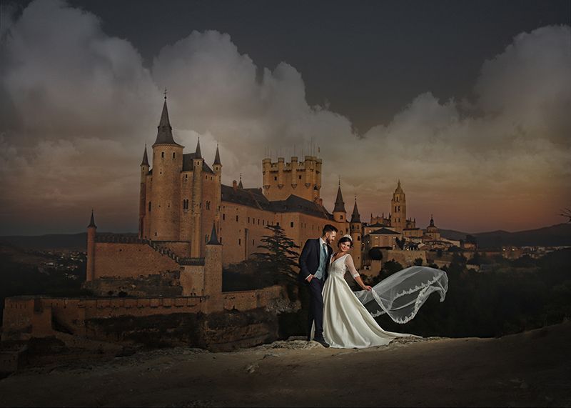 Los novios posan con un castillo de fondo al anochecer, con el velo flotando.