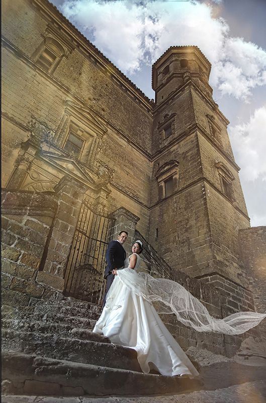 Novios en escalones de piedra frente a un alto y antiguo edificio de ladrillo. Velo ondeando. Luz del sol.