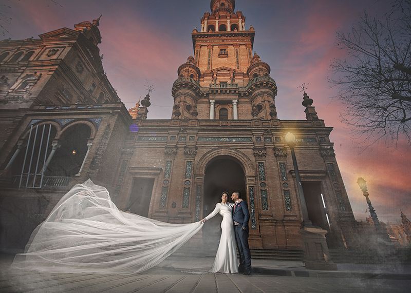 Pareja de novios en la Plaza de España, Sevilla. La novia con vestido blanco y velo largo, el novio con traje, una romántica puesta de sol.