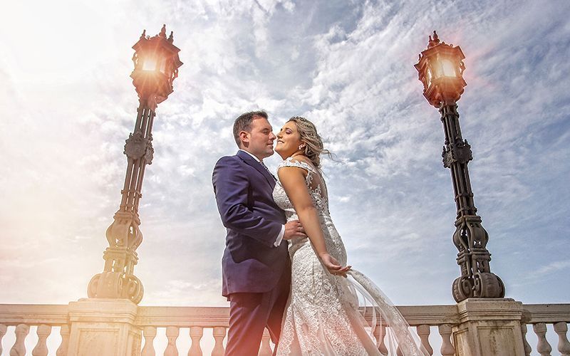 Una pareja de recién casados se abraza al aire libre, junto a farolas ornamentadas y contra un cielo nublado.