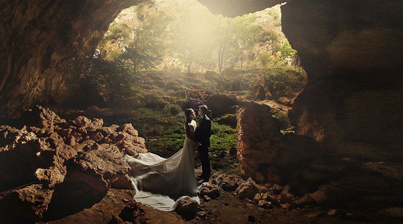 Los novios se abrazan en la entrada de una cueva iluminada por el sol. La novia lleva un vestido blanco y el novio un traje negro.